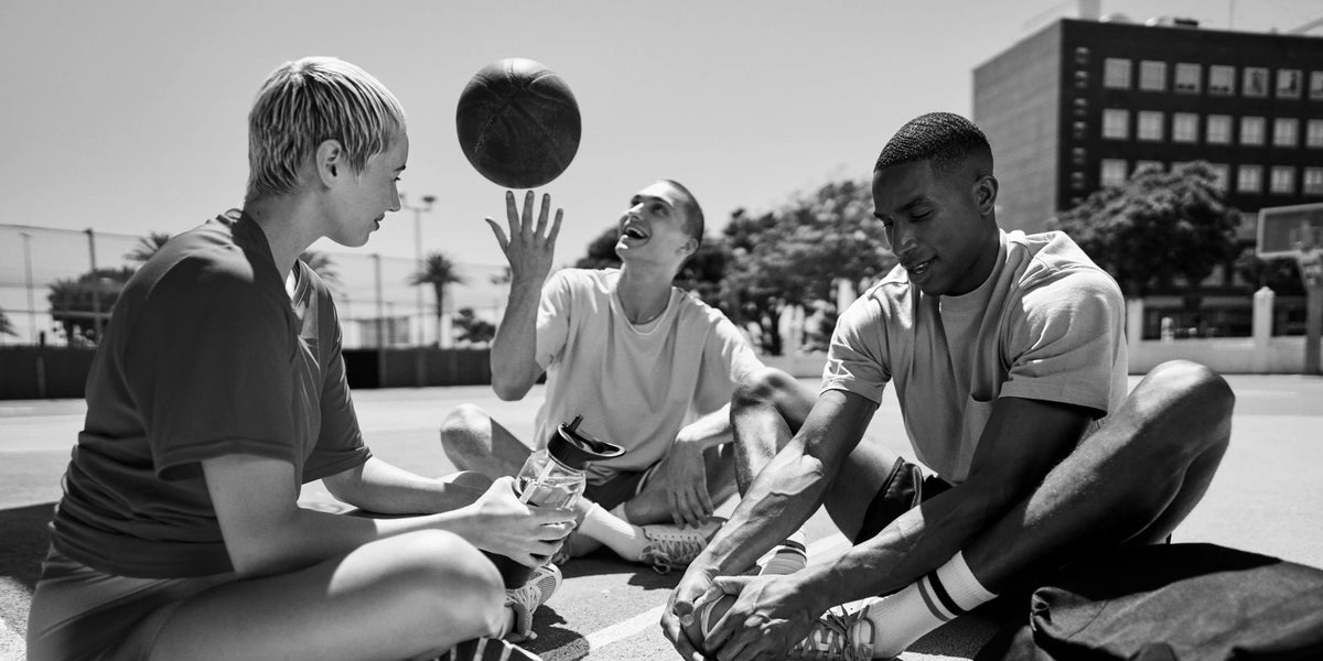 Eine Person wirft einen Basketball, eine andere dehnt sich, und die dritte hält eine Wasserflasche.